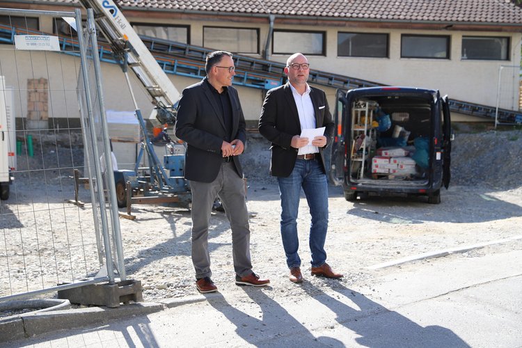 Bauprojekt der SWG Zwei Männer in formeller Kleidung stehen auf einer Baustelle vor einem Gebäude und besprechen etwas. Im Hintergrund ein Transporter mit geöffnetem Kofferraum und Baugerüste.