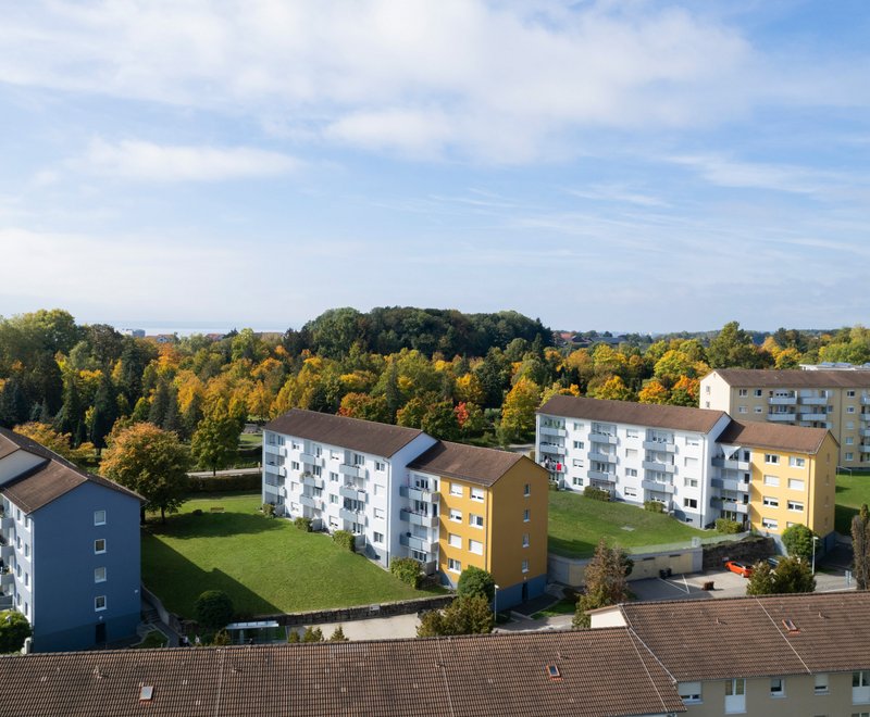 Mehrere moderne Wohngebäude Luftaufnahme mehrerer moderner Wohngebäude mit Grünflächen und Bäumen im Herbst, unter einem teilweise bewölkten Himmel.