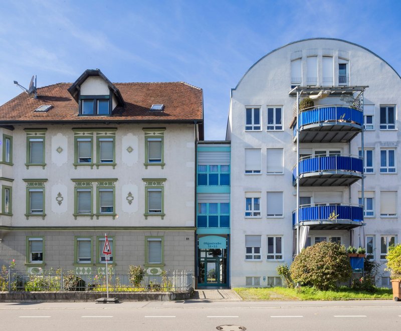 Wohngebäude der SWG Zwei benachbarte Wohngebäude, eines mit klassischer Architektur und Satteldach, das andere moderner mit abgerundetem Dach und blauen Balkonen, verbunden durch einen gläsernen Eingangsbereich. Vor den Gebäuden verläuft eine Straße.