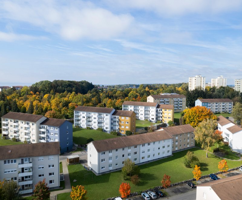 Wohngebiet der SWG Luftaufnahme eines Wohngebiets mit mehreren mehrstöckigen Wohnhäusern, umgeben von Bäumen mit herbstlichen Farben und Grünflächen, Parkplätzen und klar blauem Himmel.