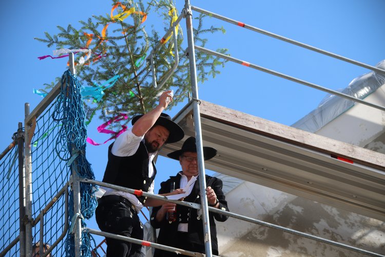 Richtfest SWG Zwei Zimmermänner in traditioneller Arbeitskleidung stehen auf einem Gerüst vor einem Bauwerk. Über ihnen ist ein mit bunten Bändern geschmückter Richtbaum befestigt, was auf ein Richtfest hinweist.