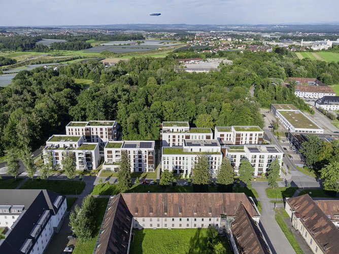Luftaufnahme eines modernen Wohnkomplexes mit mehreren mehrstöckigen Gebäuden, umgeben von vielen Bäumen und Grünflächen, in einer stadtnahen Lage mit Feldern und weiteren Gebäuden im Hintergrund.
