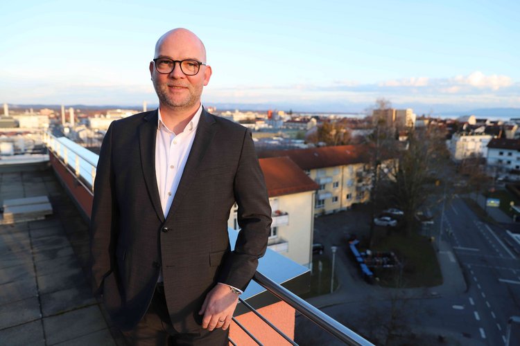 Alrik von Kolzenberg bei Bauprojekt Ein Mann in einem dunklen Anzug steht auf einem Balkon, im Hintergrund ist eine Stadtlandschaft bei Tageslicht zu sehen.