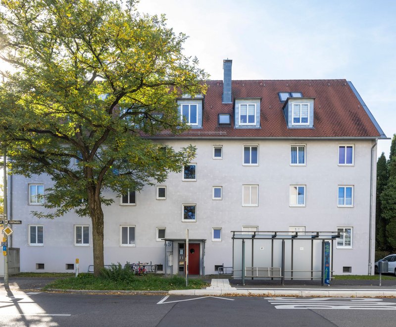 Eberhardstraße SWG Friedrichshafen Mehrstöckiges Wohnhaus mit rotem Dach und mehreren Fenstern, davor eine Bushaltestelle mit Glasüberdachung und eine große Baumkrone rechts daneben.
