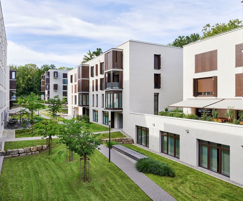 Moderne Architektur im Quartier Fallenbrunnen Moderne, mehrstöckige Wohngebäude mit großen Fenstern und Balkonen, umgeben von gepflegten Grünflächen, Bäumen und einem Gehweg zwischen den Häusern.