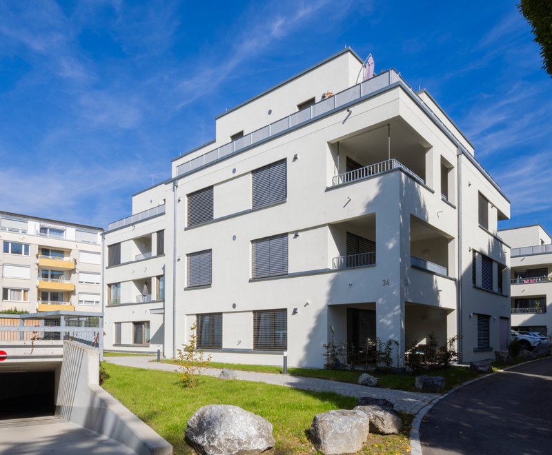 Wohnkomplex der SWG Moderner, mehrstöckiger Wohnkomplex mit weißen Fassaden, Balkonen und begrünter Umgebung unter klarem blauem Himmel. Neben dem Gebäude befindet sich eine Zufahrt zu einer Tiefgarage.